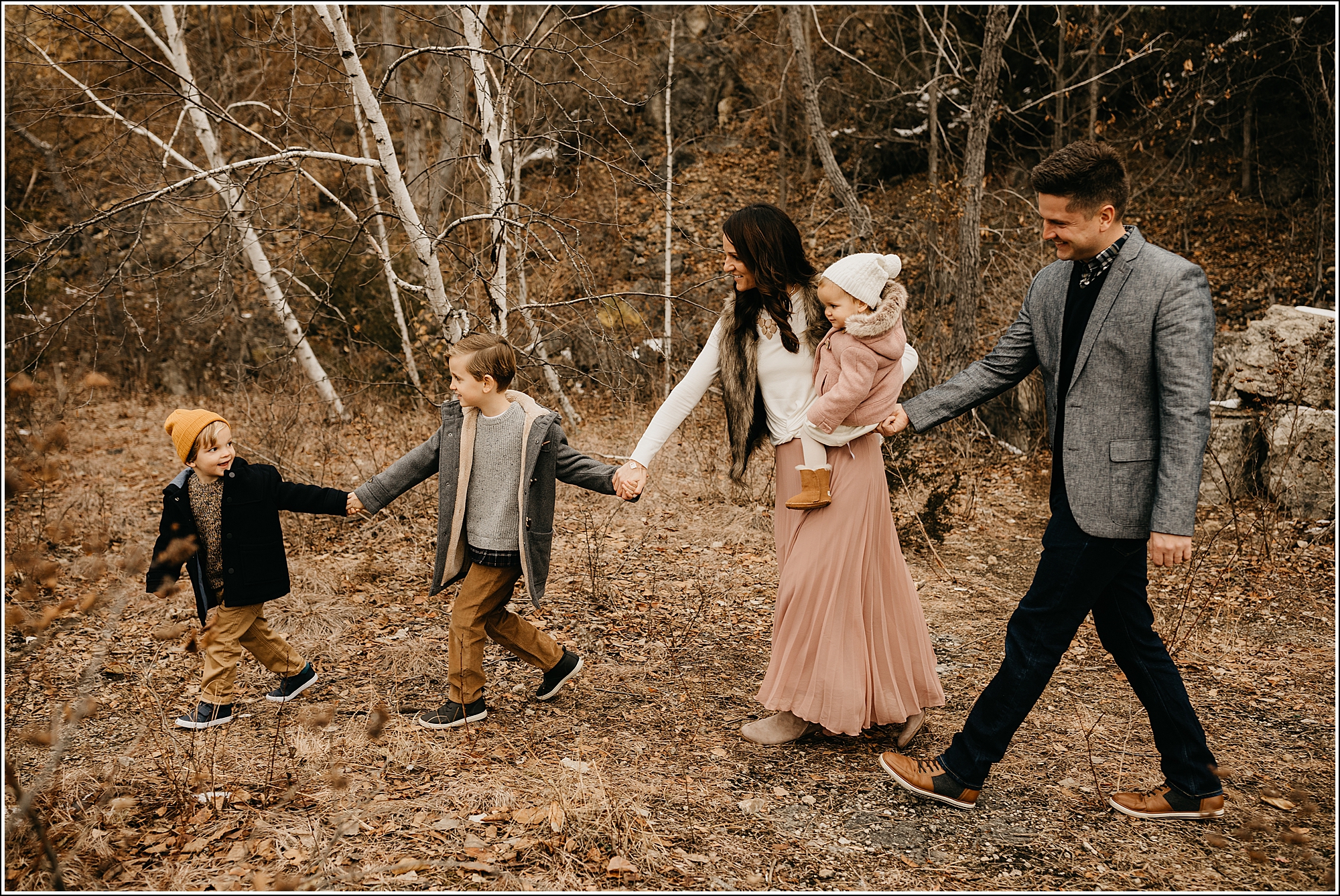 Family with great styling for family photos. Mom in long pink pleated maxi skirt. little boys in tan pants, mustard hat. dad in grey jacket, tan shoes, little girl in pink winter coat with fur, tan boots, white sweater tights